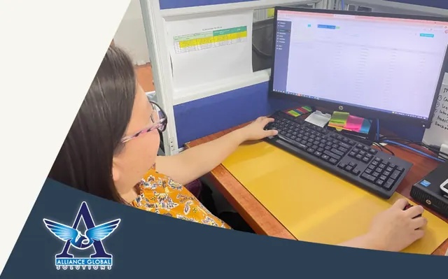 Alliance Global Solutions team member working at a cubicle computer with spreadsheet software and sticky notes on the monitor